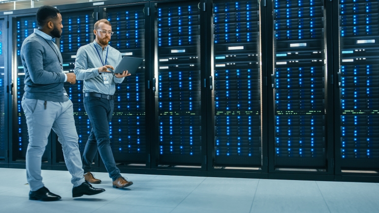 IT Technician with a Laptop Computer and Black Male Engineer Colleague are Talking in Data Center while Walking Next to Server Racks. Running Diagnostics or Doing Maintenance Work.