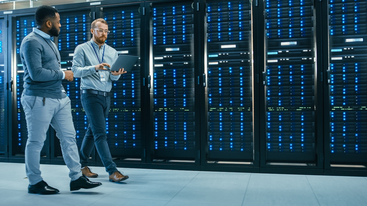 IT Technician with a Laptop Computer and Black Male Engineer Colleague are Talking in Data Center while Walking Next to Server Racks. Running Diagnostics or Doing Maintenance Work.